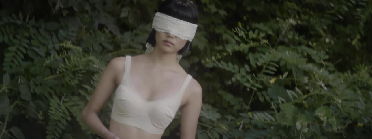 A woman wearing her undergarments and a lace blindfold stands in a garden holding a peach