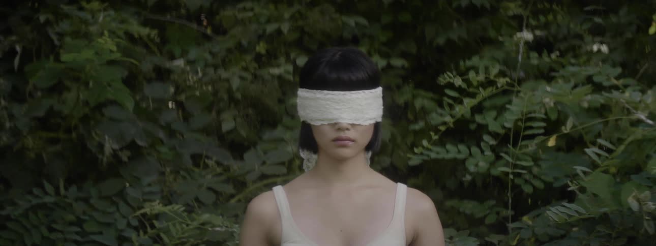 An asian woman wearing a blindfold stands in front of a bushes