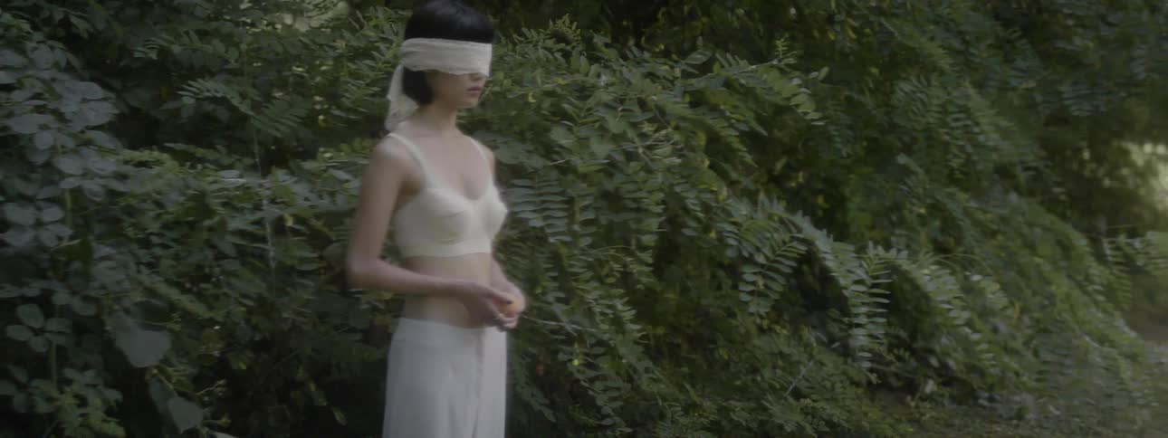 A woman in a blindfold standing in the shadows of the trees at the edge of a field while clasping a fruit between her hands