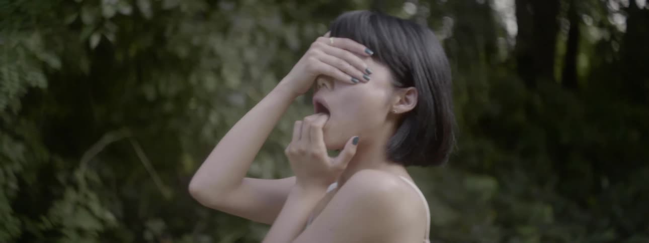 Young woman with hand over eyes and hand holding mouth open in front of trees