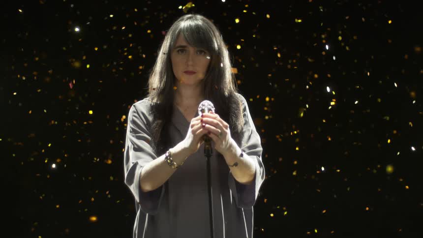 Woman stands at microphone with gold confetti falling around her
