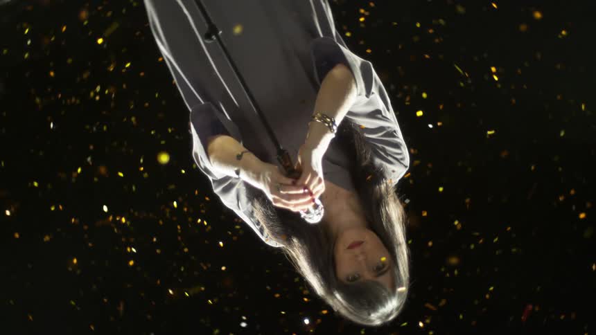 Gold and silver sparks and flecks glittering around a woman holding a microphone on a stand while staring emotionlessly into the spinning camera