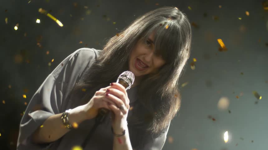 Woman singing into microphone surrounded by falling gold confetti