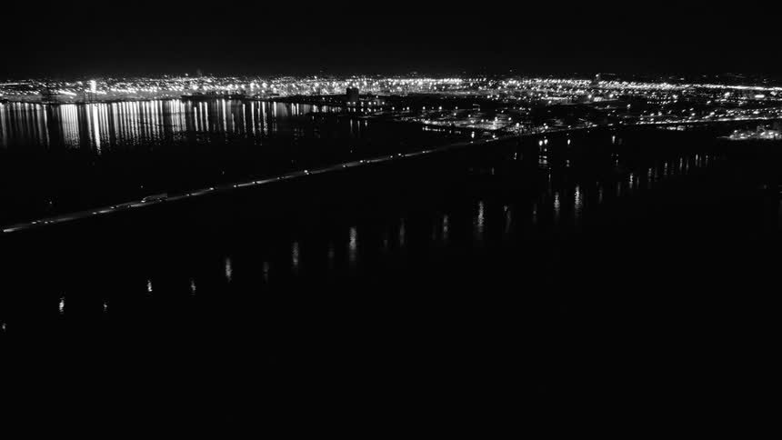 Black and white film of traffic traveling on long bridge over river in front of city at night
