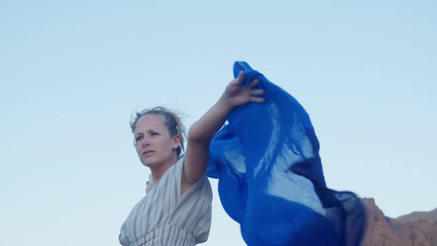 A woman stands in a desert with her arms raised over her head and the material she is holding blowing in the wind