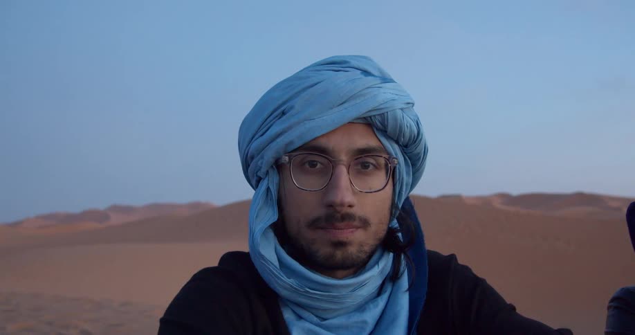A man in a blue turban poses in a desert and gives a wry smile