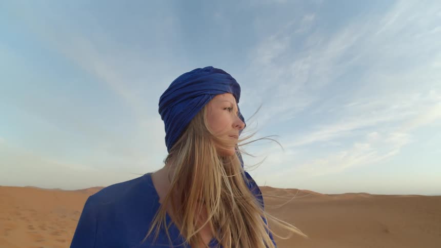 A white woman in a blue cloak and head covering stands in the desert looking around