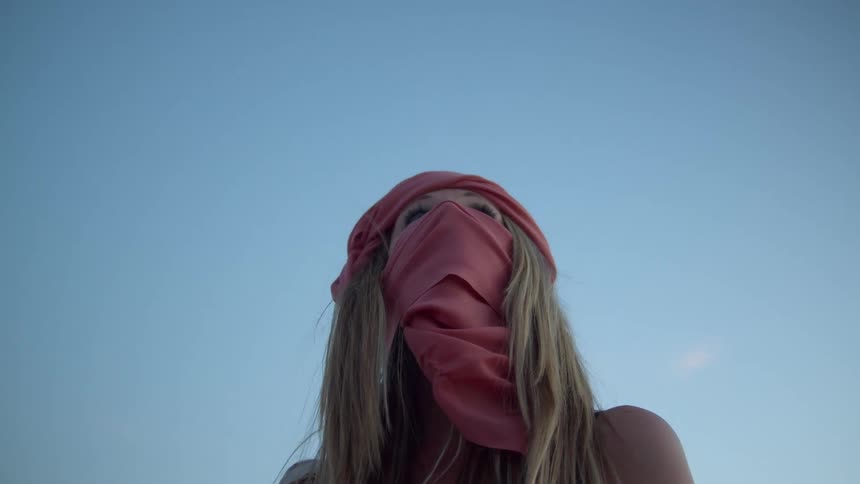 A woman with her head and face wrapped with a pink clothing looking up at the sky before lowering her gaze to look into the camera