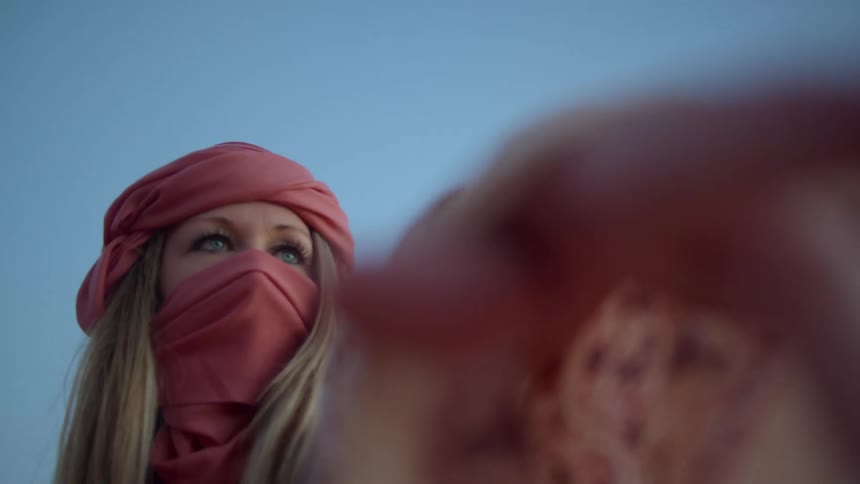 A white woman wearing a turban and veil stands outdoors and points her arm and hand