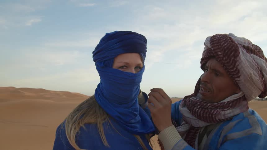 Man wearing a turban adjusts the face covering of a woman in the desert