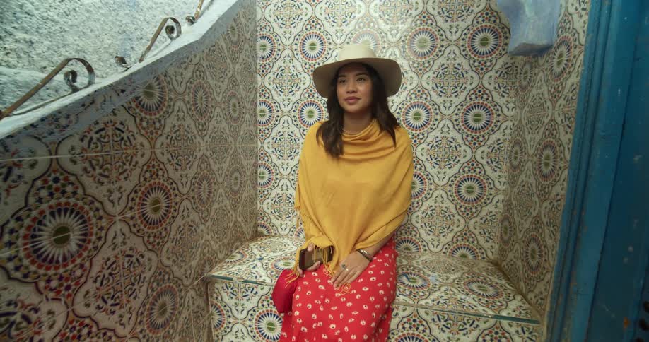 An asian woman in a hat and colorful garb sits on a bench covered in decorative tiles