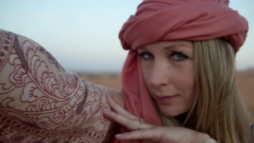 A white woman with a turban stands in a desert with her arm and hand extended