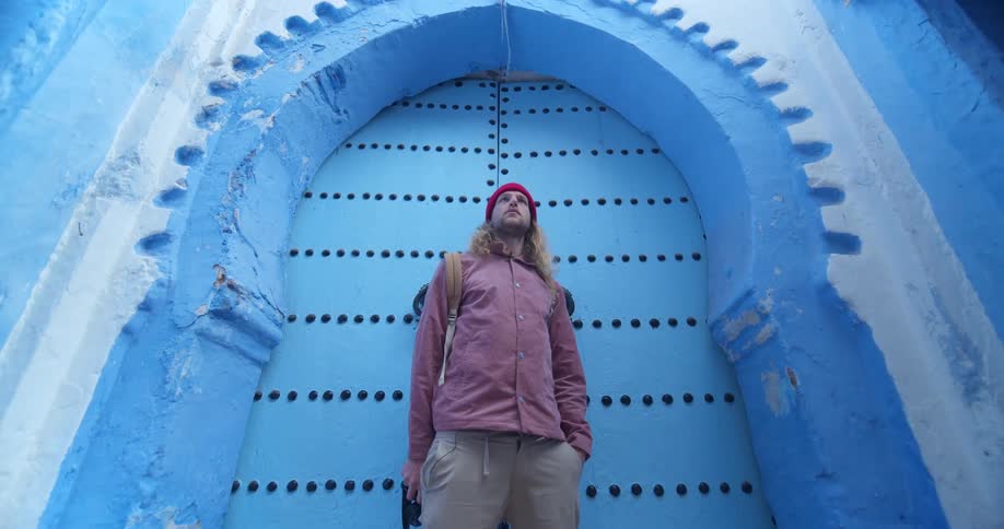 A man with long blond hair poses in front of a blue abode building with an arched doorway