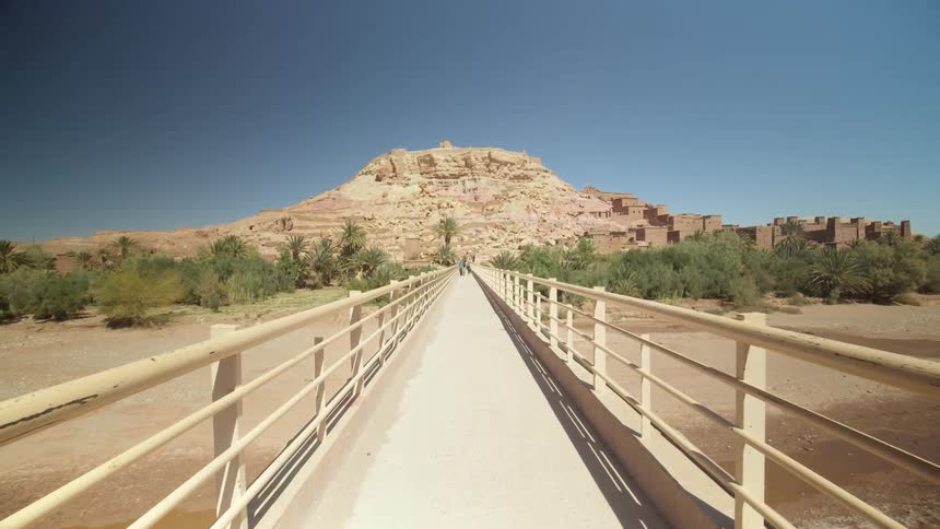 A walking bridge over a river leads to a mountain in the desert