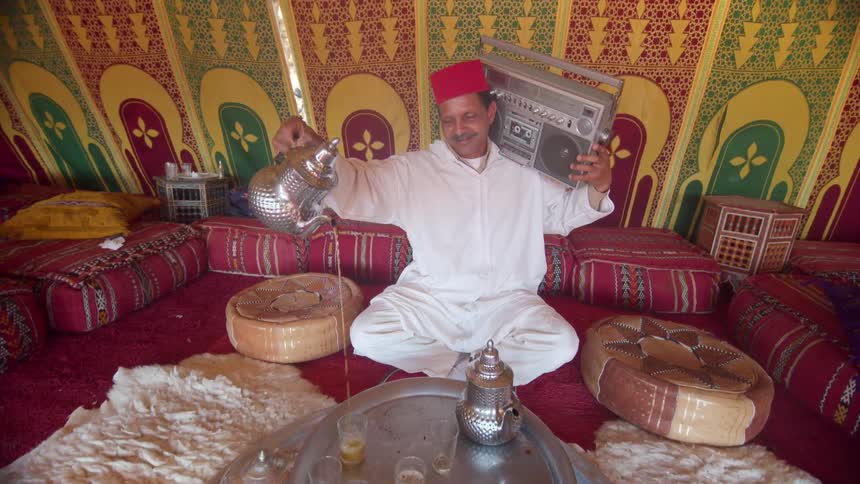 A man sits in a large colorful tent with a boombox on his shoulder pouring tea into a cup from a silver teapot