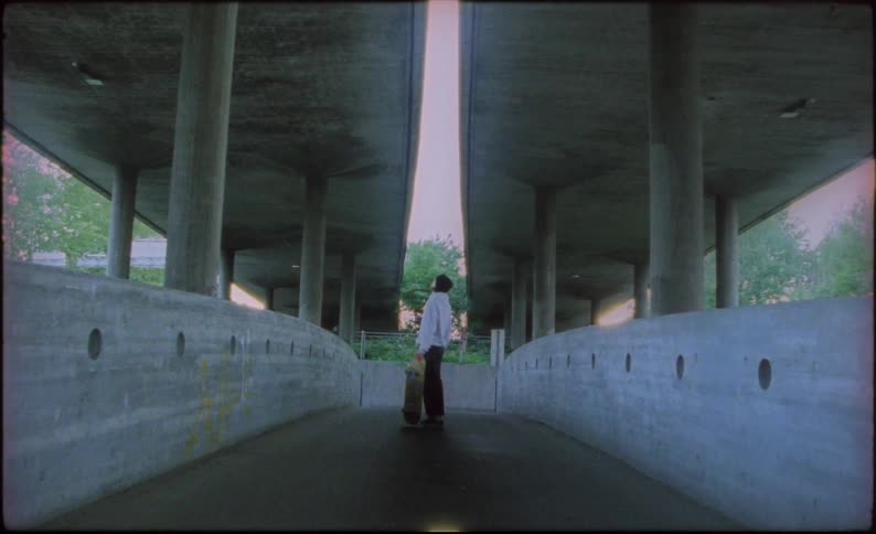 A boy holds his skateboard in a walk way and looks up