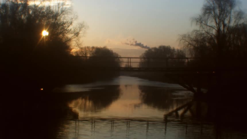 The fence along the bridge and trees are reflected in the water surface at sunset