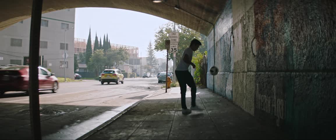 A man kicking a football underneath a tunnel against a wall with a street with vehicles behind him