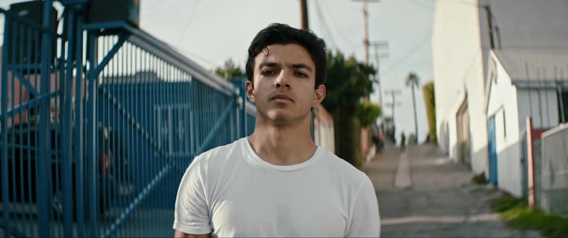 A man stands on a narrow street next to a fence staring straight ahead