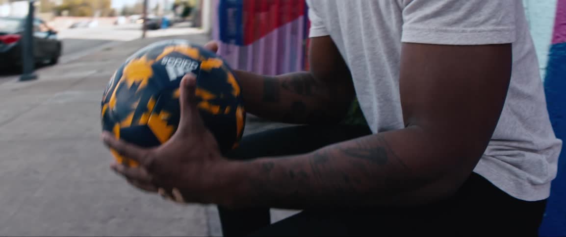 A man sits on a city bench tossing a soccer style ball to himself