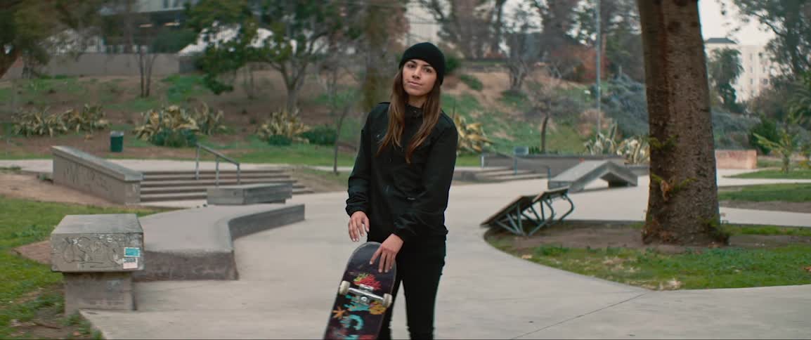 Woman wearing jacket and beanie stands in a park with a skateboard on her hand