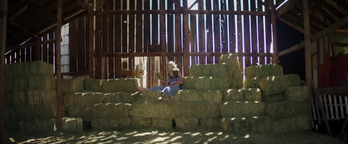 Mother has her arm around her daughter as they sit on top of bales of hay inside of a barn reading a book with one another