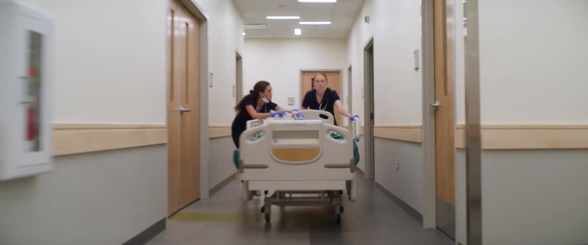 Wooden double doors open quickly as two female nurses run through the hallway while pushing a patient in a hospital bed