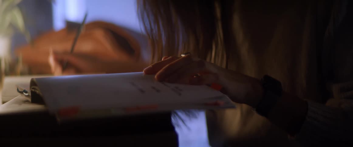 A female sits at a desk with a book and pencil leaning forward