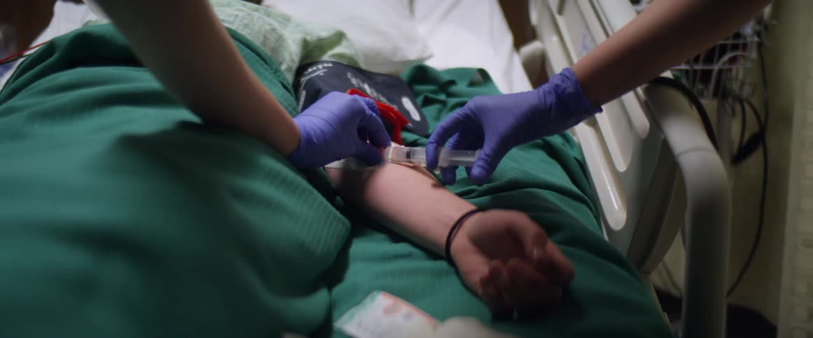 Female nurse quickly removes the cap off of a syringe and attaches it to a needle in the patients arm who is lying unconscious in a hospital bed as another nurse stand by to help out