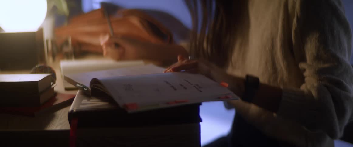 A woman sitting on a desk writing in a notebook then looks at her watch and gets up and leaves