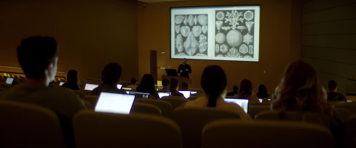 Students sit in a lecture hall with their laptops watching an instructor give a lecture
