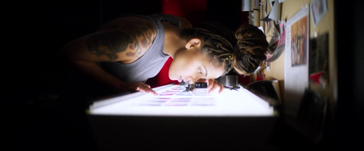 Woman examines film on light table with photo covered bulletin board in background