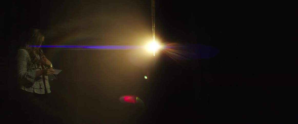 Light flares through a crack in a stage curtain as a white woman stands to the side preparing to speak with notecards