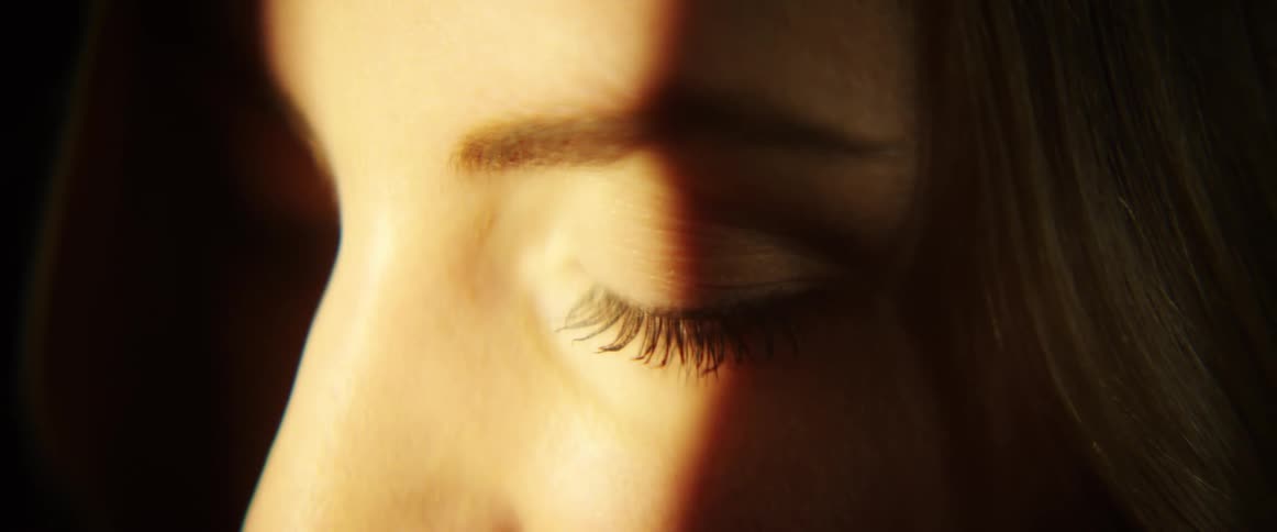 A white woman stands in a band of light and closes and opens her eye