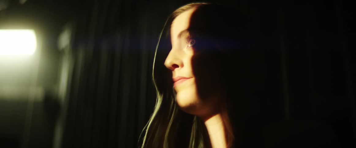 A white woman stands on a stage and looks at the light coming through a crack in the curtain