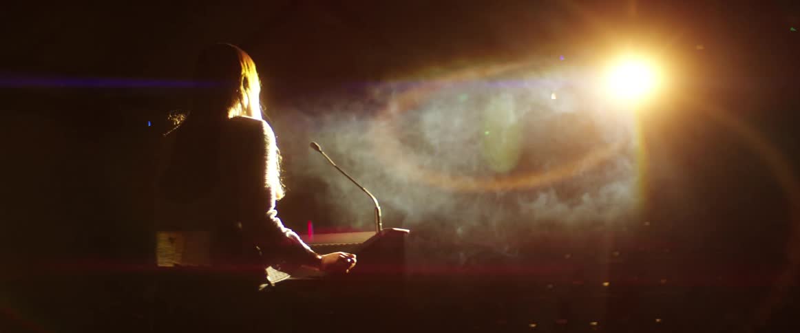 A white woman walks up to a podium illuminated by a bright spotlight