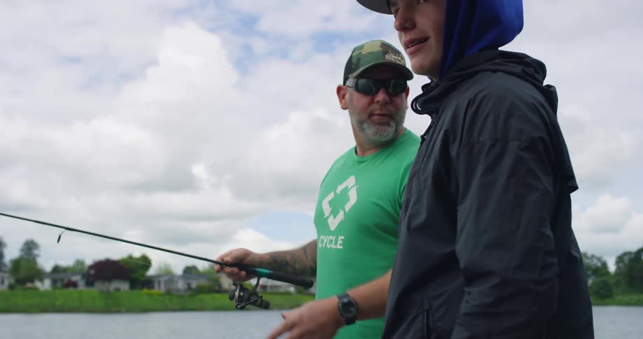 A father and son stand near a lake with a fishing pole and talk to each other