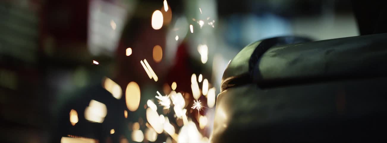 Two men inside a workshop wearing safety glasses begin to weld metal and sparks begin to fly