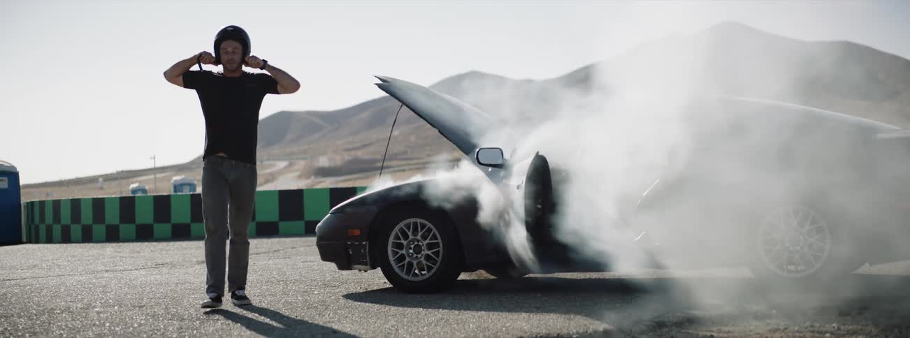 A race car driver looking at the smoking engine of his vehicle in frustration before removing his helmet and walking away