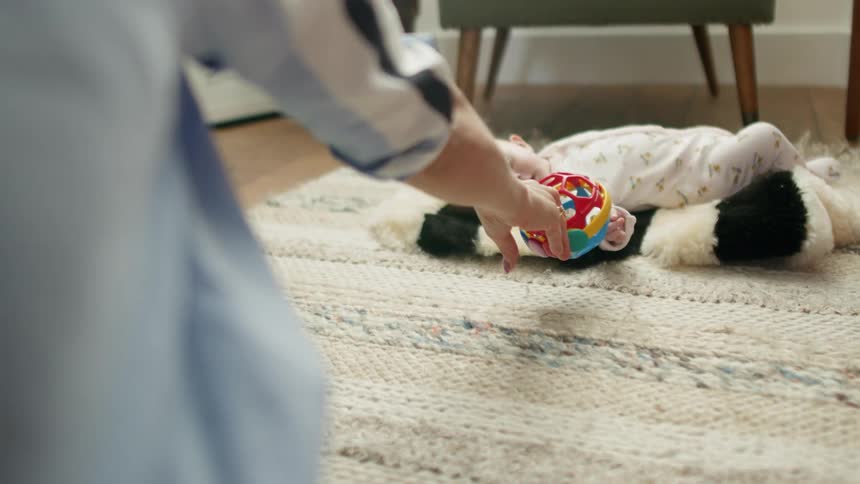 Baby laying over a rug on the floor plays with a toy with his mother