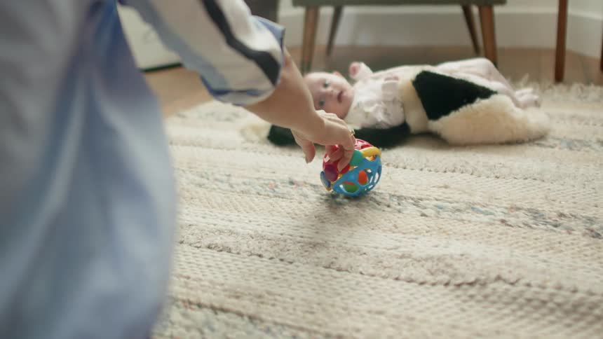 Baby laying on the floor as her mother rolls a colorful toy towards her and tries to get the baby to grab ahold of the toy