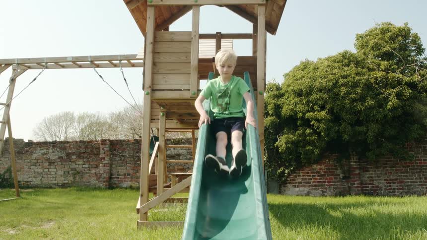 Boy slides down a sliding board attached to a wood swing set and reaches out with both hands to catch himself in the grass