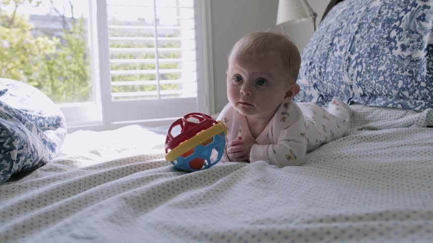 Baby wearing lies down on stomach on bed near open window showing trees