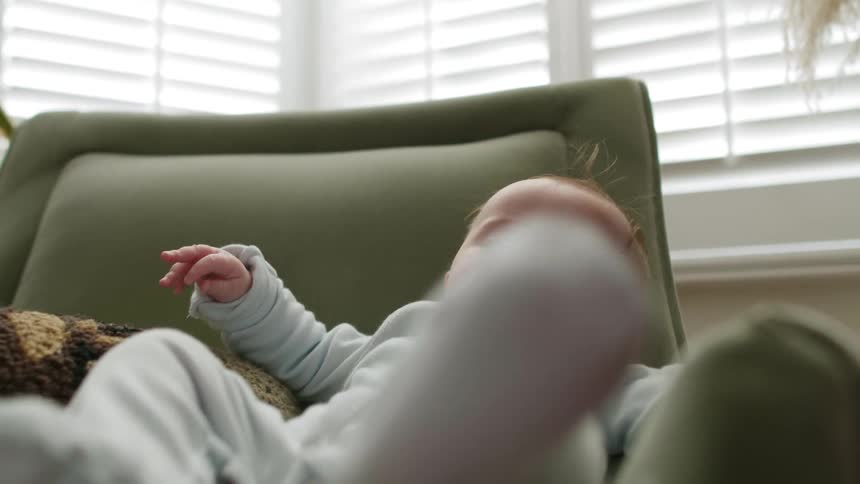 Baby kicks its legs as it sits in a chair in front of a window