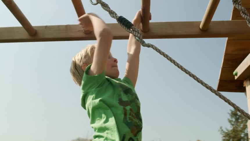 A white kid swings across monkey bars to an enclosed wooden structure
