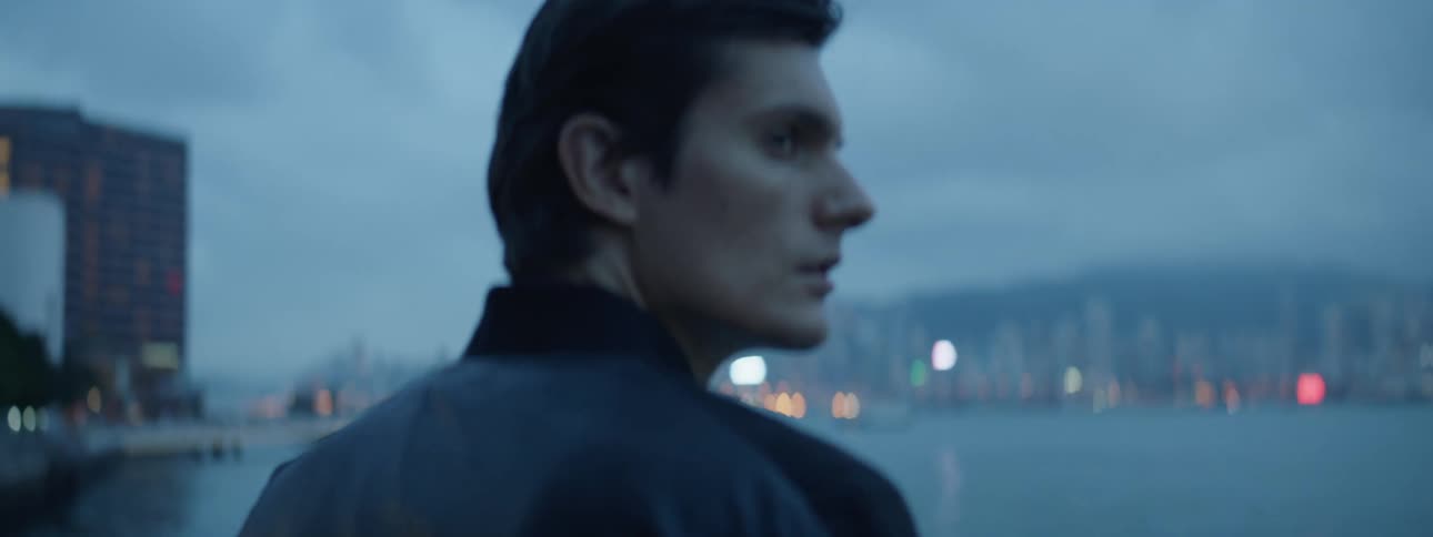 Man stands at waterfront with city in distance at dusk