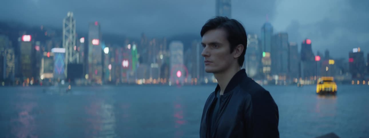 A white man stands on a deck next to a bay near a city and stares off into the distance