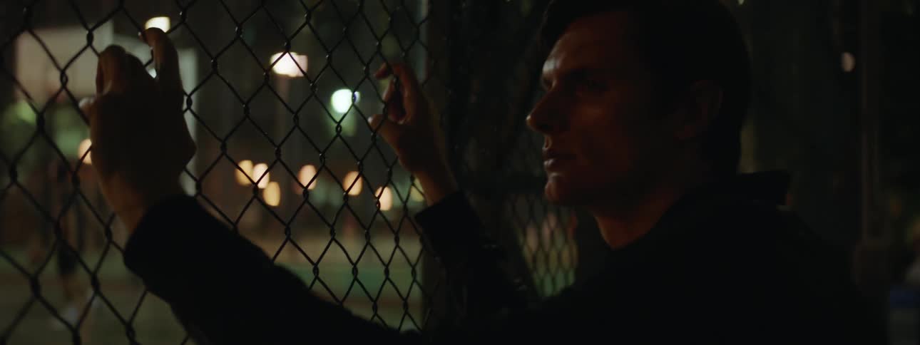 Man holding onto wire fence watches the people playing basketball at night