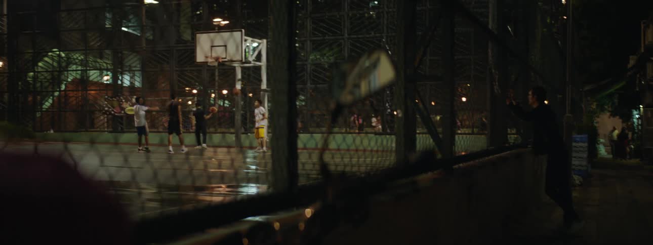 A man watching another a group of men play basketball on an outdoor basketball court at night