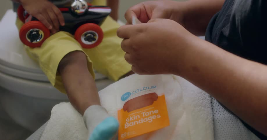 A black woman takes a bandage out of a package for a toddler who is sitting on a toilet holding a toy car